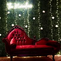 Romantic red velvet chaise lounge set against a dark backdrop with hanging string lights, creating a moody and elegant atmosphere.