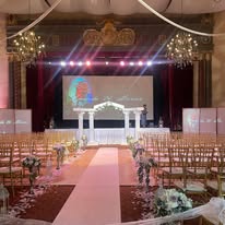 A wedding ceremony setup with rows of chairs leading to an altar under a grand chandelier, with soft lighting and a colorful projection on the backdrop.