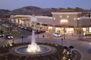 Outdoor plaza at dusk featuring a central illuminated fountain, surrounding walkways, and a warmly lit event venue with guests gathered around.